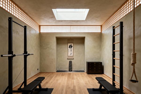 A minimalist home gym with light oak flooring and warm beige plaster walls features a central tokonoma alcove displaying a vertical calligraphy scroll above a standing stone set on black pebbles. Perimeter shoji-style clerestory screens and a square skylight bathe the room in soft daylight, while a black squat rack with barbell and bench sits left, Swedish wall bars and a climbing rope hang right, and a low dark tansu chest rests near the rear wall.