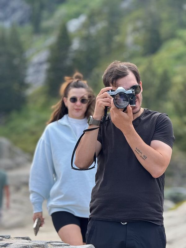 A man is taking a photo with a camera while a woman walks behind him.