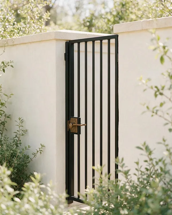 A simple transitional-style exterior gate framed by smooth white stucco walls and topped with flat coping. The black metal gate has vertical bars and a brass latch, while soft green shrubs and blurred foliage surround the entrance in warm daylight.