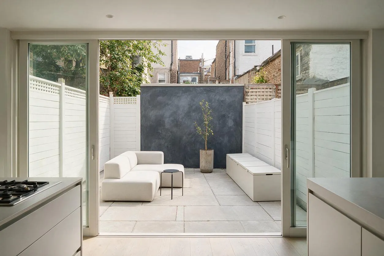 A minimalist backyard courtyard viewed through wide sliding glass doors, featuring white fence panels, pale stone paving, a low white sectional sofa, a small round side table, a long storage bench, and a dark charcoal feature wall with a potted tree. Soft natural light and restrained materials create a calm, modern outdoor retreat.