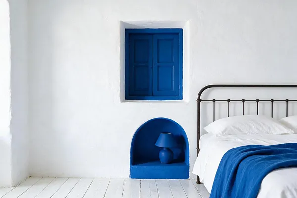 A minimalist Mediterranean-style studio bedroom with whitewashed walls and pale painted floorboards. A dark metal bed with crisp white bedding and a cobalt blue throw sits beside a small arched niche holding a matching blue lamp, while a blue shuttered wall opening adds a bold focal point.