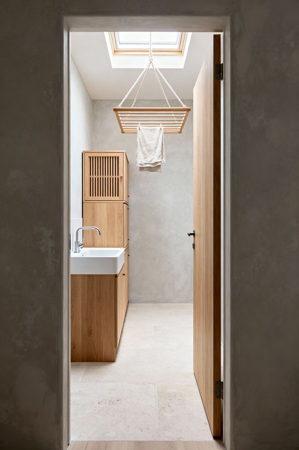 A narrow Scandinavian-style laundry room with light troweled plaster walls and large pale stone tiles, seen through an open oak door. Oak cabinets line the left side with a white rectangular ceramic sink and chrome tap, while a slatted wooden ceiling airer hangs from thick ropes beneath a skylight, drying a single linen towel. The soft neutral palette highlights natural materials, clean lines, and ample daylight.