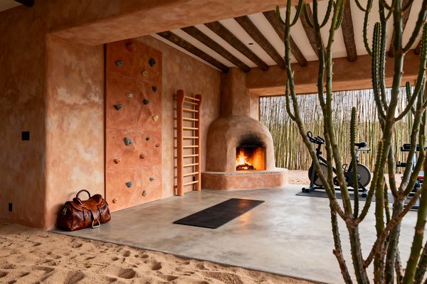 A Southwestern-style home gym clad in sunbaked terracotta plaster features exposed dark wood ceiling beams and a rounded adobe kiva fireplace with a live fire. A terracotta climbing wall with multicolored holds and a wooden Swedish ladder sit beside a black yoga mat on polished concrete that transitions to an indoor sand border; stationary bikes rest near an open side screened by vertical cane, with desert cacti and warm natural light beyond.