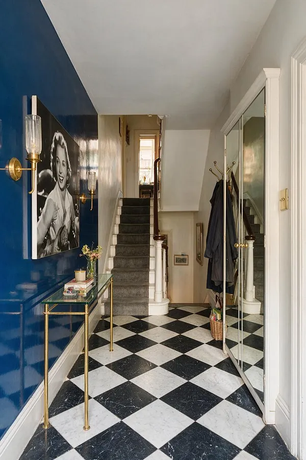 A Hollywood Regency entryway with a glossy cobalt blue accent wall, black-and-white checkered marble floor, and a large mirrored closet door. Brass wall sconces, a slim glass-top console, and a black-and-white portrait add glamour beside a carpeted staircase.