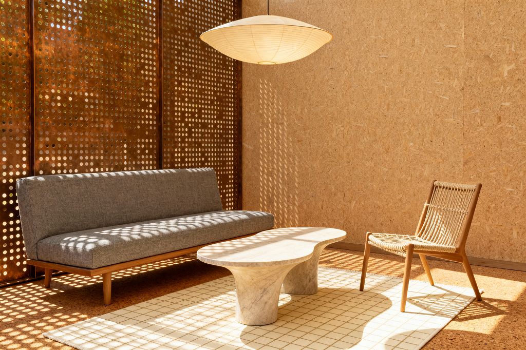 A sunlit modern living room with a bronze-toned perforated metal screen casting circular shadows across OSB wood walls and cork flooring. A low gray upholstered sofa with an oak base faces a sculptural pale travertine coffee table on a cream grid rug, paired with a natural cord lounge chair. An oversized saucer-shaped paper lantern hangs overhead, creating a warm, minimal atmosphere with clean lines and a restrained neutral palette.
