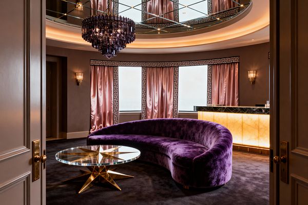 Opulent living room with a curved purple velvet sofa on a dark carpet, a round glass coffee table with a gold starburst base, and a backlit onyx-front bar topped in black marble. Blush satin curtains with black Greek key trim line bay windows, while ribbed glass sconces and a dark amethyst crystal chandelier glow beneath a mirrored coffered ceiling with cove lighting.