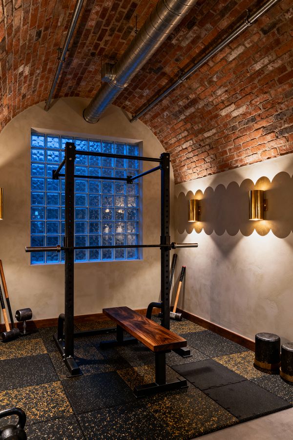 A home gym set beneath a barrel‑vaulted red‑brick ceiling with exposed galvanized ductwork and metal conduits. A black steel power rack stands over speckled black rubber tiles, paired with a narrow walnut bench. A glass‑block window casts cool blue light, while two cylindrical brass sconces wash warm light across a scalloped accent band on plastered walls. Kettlebell, dumbbell, and mace-style tools rest along the perimeter.