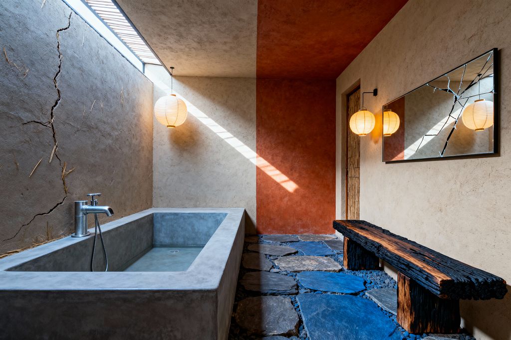 A wabi-sabi bathroom features a rectangular cast-concrete soaking tub beneath a narrow skylight, sending a diagonal beam of light across textured plaster walls. Natural lime plaster with visible straw and fissures contrasts a terracotta wall and ceiling, while round paper lanterns, a cracked rectangular mirror, irregular blue‑gray stone pavers set in black pebbles, a charred timber bench, a bamboo door panel, and a simple brushed-metal faucet complete the pared-back scene.