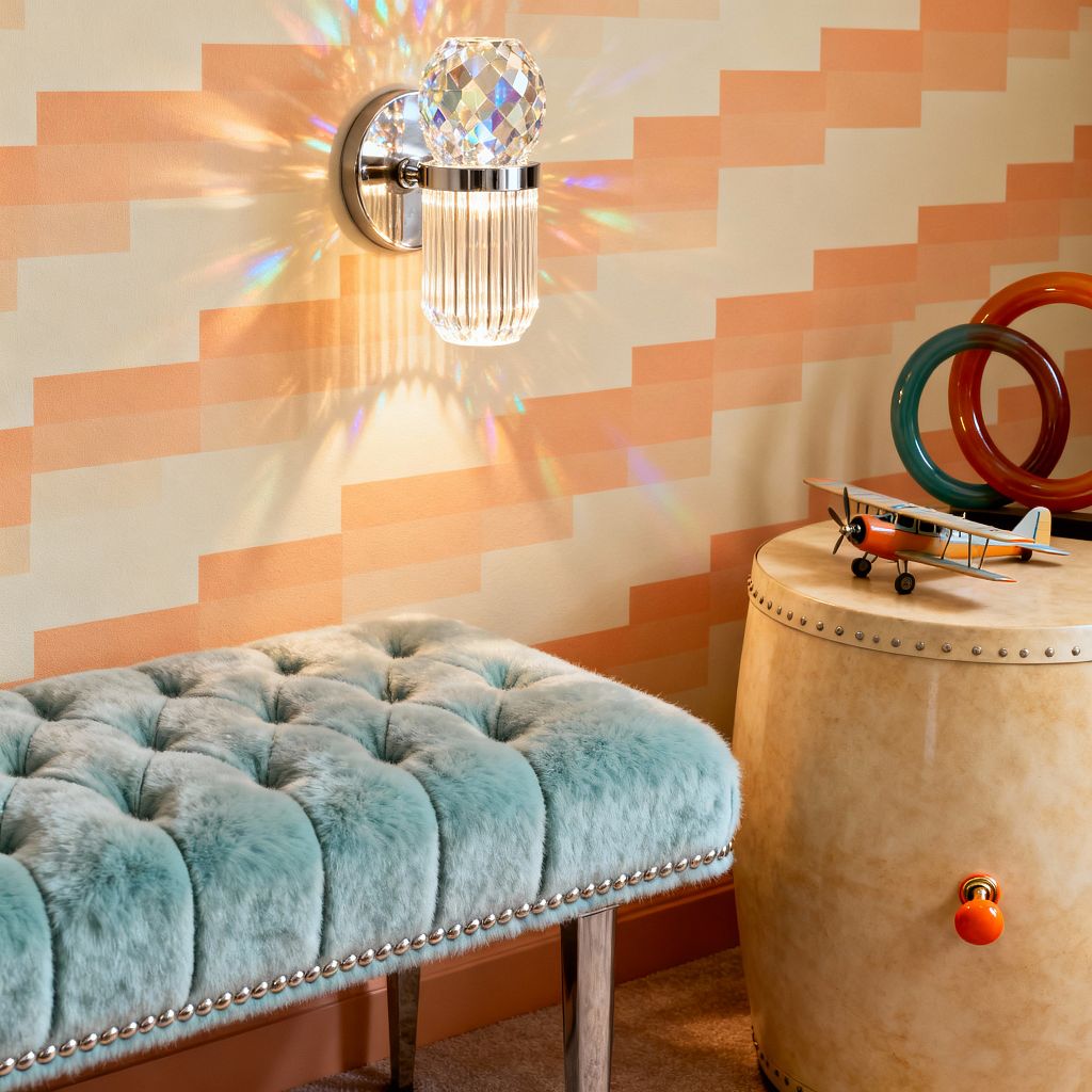 Close-up of a kids’ room corner with a polished nickel wall sconce featuring a faceted crystal cap and ribbed glass shade that casts rainbow reflections across peach-and-cream stepped wallpaper. Below is a seafoam tufted faux-fur bench with nailhead trim and metal legs. To the right, a parchment-finish cylindrical side table with studded edges holds colorful acrylic rings and a small model airplane; beige carpet and a terracotta baseboard complete the scene.