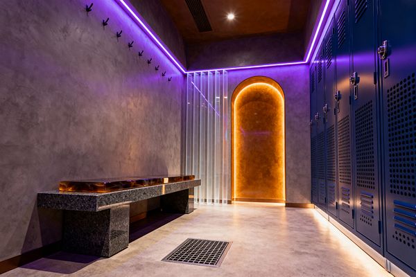 A cyberpunk-style mudroom features deep-blue perforated steel lockers on the right, gray concrete walls and a polished concrete floor, and violet LED strips tracing the ceiling edges. Opposite, a granite bench with a translucent amber resin seat sits below a row of black hooks, while a warm backlit arched niche and ribbed clear divider add layered lighting; toe-kick illumination and a central floor grate emphasize the industrial, easy-clean layout.