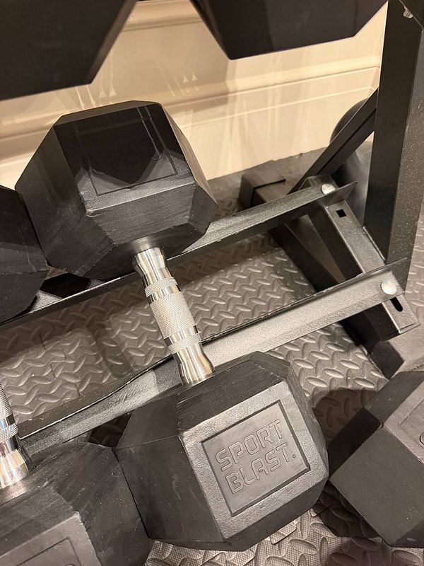Close-up of a broken gym dumbbell rack with a dumbbell sliding off, showing twisted steel and the 'SPORT BLAST' logo on the dumbbell.