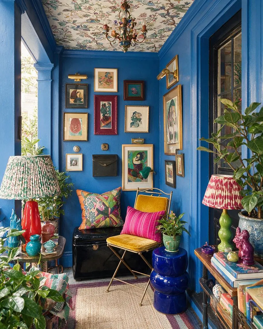 A vividly decorated front porch with bright blue walls and ceiling, a floral wallpapered ceiling, and a cluster of framed artwork arranged salon-style on the back wall. The space includes colorful patterned lamps, a mustard chair with pink and multicolor pillows, glossy blue and red accents, potted plants, and layered rugs, creating a lively maximalist look.