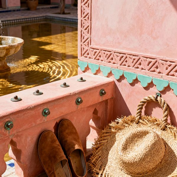 A blush pink tadelakt niche with lattice-carved trim and mint-green scalloped zellige tiles forms a Moroccan-inspired mudroom ledge. Verdigris brass studs line the edge, while tan suede slippers and a woven palm straw hat rest on the seat; in the background, a shallow courtyard fountain with a mosaic border catches sunlight, sending rippling honey reflections across the rosy plaster for a warm, handcrafted arrival moment.