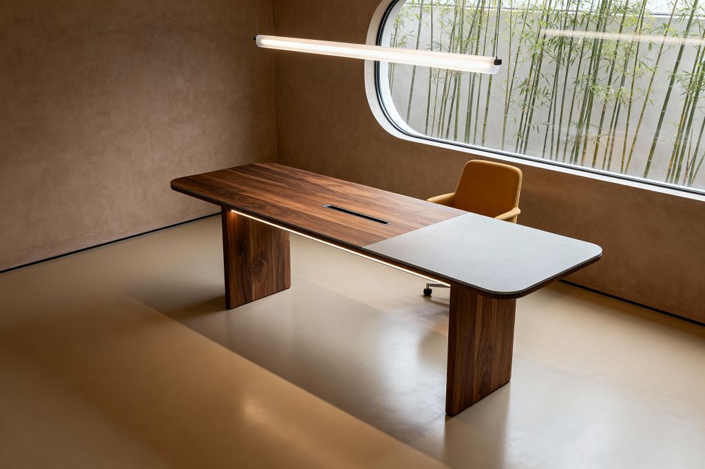 A minimalist home office with a long walnut desk featuring a light gray stone inset and two block legs, underlit by a slim LED strip. A linear tubular pendant hangs above the desk. A mustard upholstered swivel chair sits by a large window with rounded corners, showing bamboo outside. The room has taupe plaster walls and a smooth beige resin floor, with a central cable slot on the desktop and black window framing.