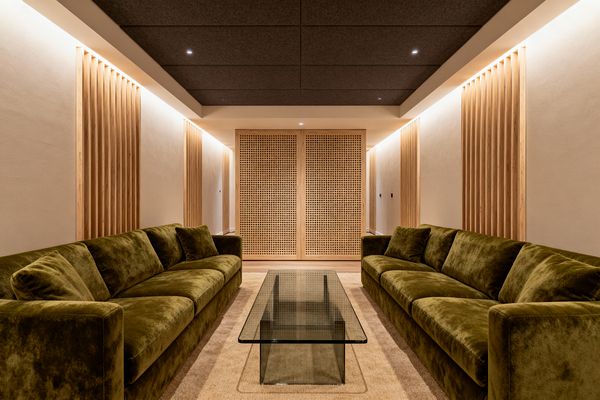 A symmetrical media room with two long olive-green velvet sofas facing across a clear glass coffee table on a beige carpet. Light, warm-white walls are accented with vertical natural-wood slat panels and a perforated wood screen at the back, while soft perimeter cove lighting and small recessed downlights illuminate a charcoal panelled ceiling, creating a warm, quiet atmosphere suited to relaxed viewing.