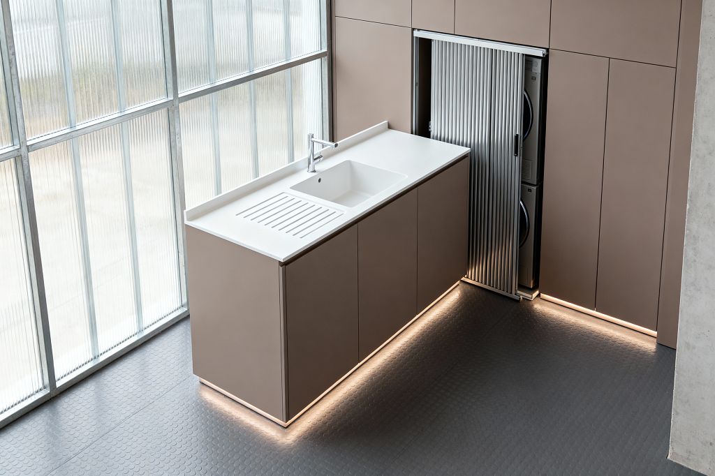 A minimalist laundry room with a floating taupe cabinet run topped by a white solid-surface counter and an integrated sink with slotted drainboard, centered by a chrome single-lever faucet. Tall, handleless taupe cupboards line the wall; a ribbed aluminum tambour door slides open to reveal a stacked washer and dryer. LED toe-kick lighting highlights the base, while charcoal rubber coin flooring and full-height ribbed glass windows deliver soft, diffused daylight.