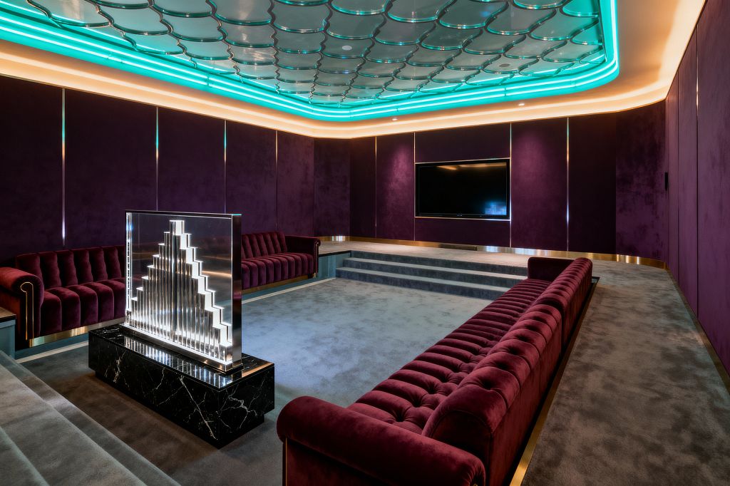 A luxurious media room features deep plum velvet wall panels and matching channel‑tufted sofas around a sunken, tiered gray carpet facing a built‑in flat screen. At center sits an illuminated glass and chrome Art Deco divider on a black marble base, while a scalloped glossy ceiling with turquoise cove lighting and brass accents along walls and sofa bases create a glowing, symmetrical, theater‑like setting.
