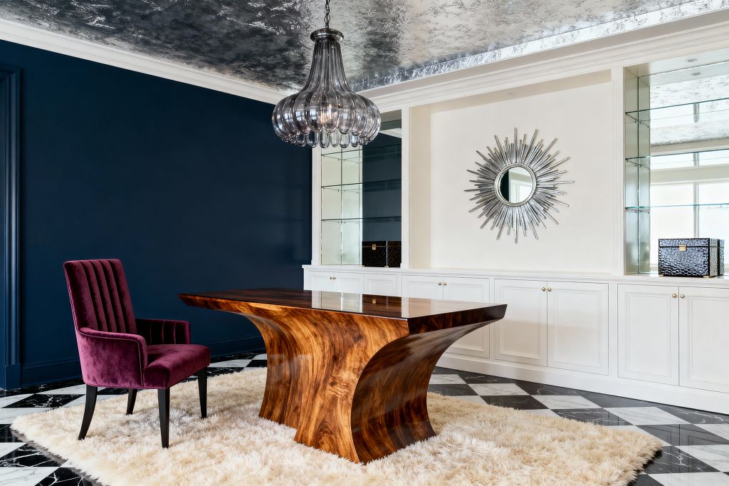 Home office with a sculptural, high-gloss walnut desk centered on a cream shag rug over black-and-white marble tiles. To the left sits a plum velvet, channel-tufted chair against a deep navy wall; to the right, white built-in cabinetry with brass knobs, mirrored shelves, and a silver sunburst mirror. A smoky glass chandelier hangs from a silver-leaf ceiling, reflecting daylight and adding glamorous sparkle throughout the room.