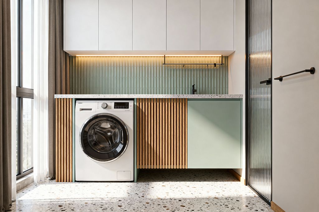 A compact contemporary laundry features a front-loading washer beneath a speckled terrazzo countertop, with white flat-panel wall cabinets above. The backsplash is sage green vertical fluted tile lit by a warm LED strip; base cabinets combine a mint-green slab door and a slatted oak panel, with a matte-black faucet and hanging rail, terrazzo flooring, a tall window with layered curtains to the left, and a reeded-glass door with black frame to the right.