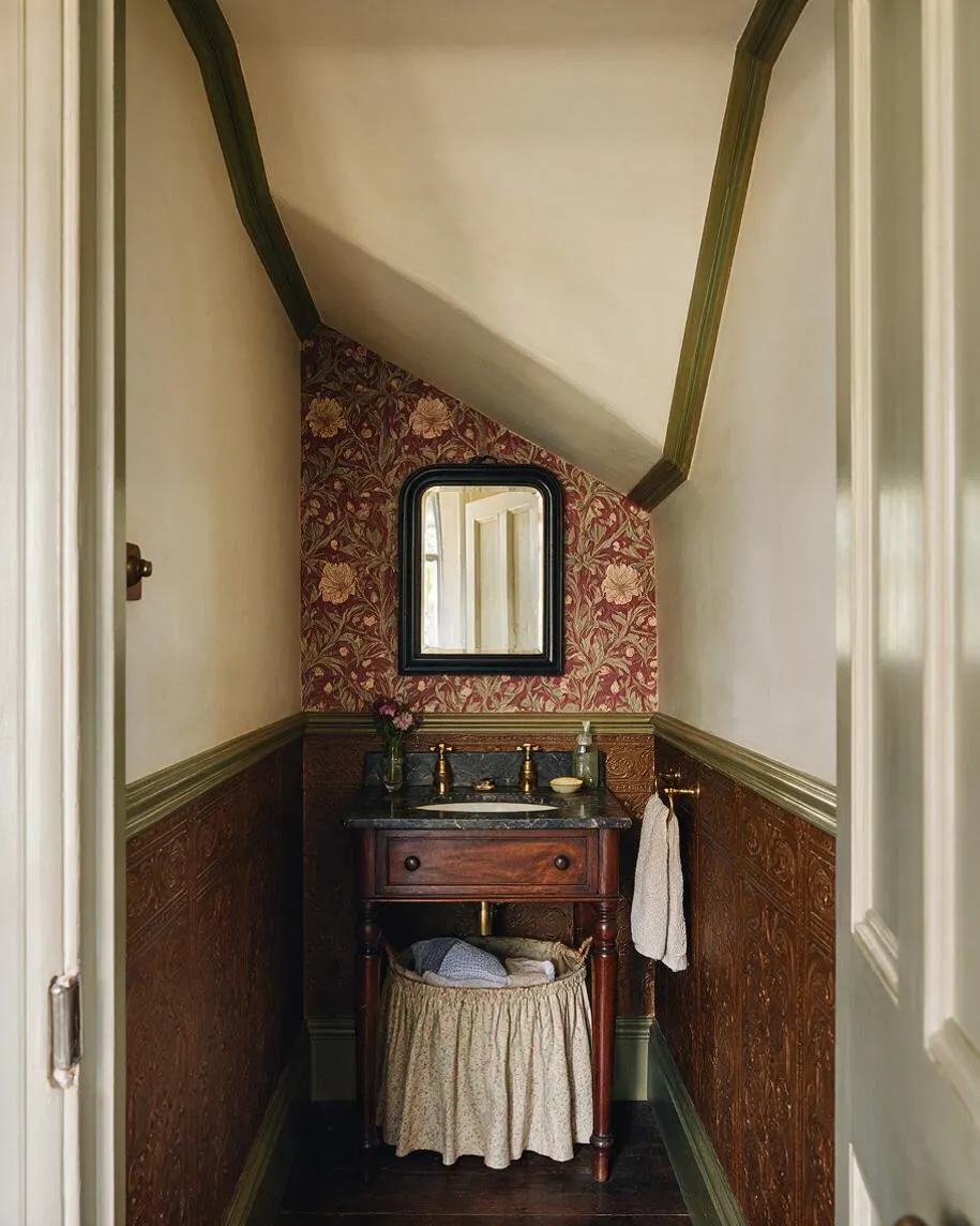 A narrow Victorian-style powder room with sloped ceilings, floral wallpaper on the far wall, and dark wood wainscoting below. A small wooden vanity with a black stone top, brass faucet, framed mirror, and woven laundry basket creates a warm, traditional look.