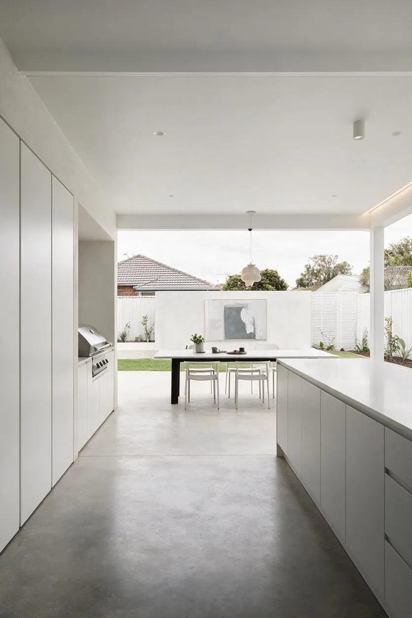 A minimalist backyard patio seen from an indoor-outdoor kitchen, featuring smooth concrete flooring, white built-in cabinetry, and a large open covered ceiling. At the back, a simple dining table sits beside a white fence, a narrow strip of lawn, and sparse planting, with a hanging pendant light and muted artwork adding subtle detail.