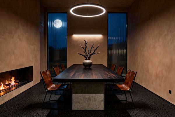 A contemporary dining room with a long dark-wood table on a concrete pedestal, surrounded by six cognac leather sling chairs on black steel frames. A glowing circular pendant and slim wall light illuminate taupe plaster walls, a linear recessed fireplace on the left, and two tall fluted-glass windows showing a night sky with the moon; a ceramic vase with sculptural branches sits centered on the table above a dark terrazzo floor.