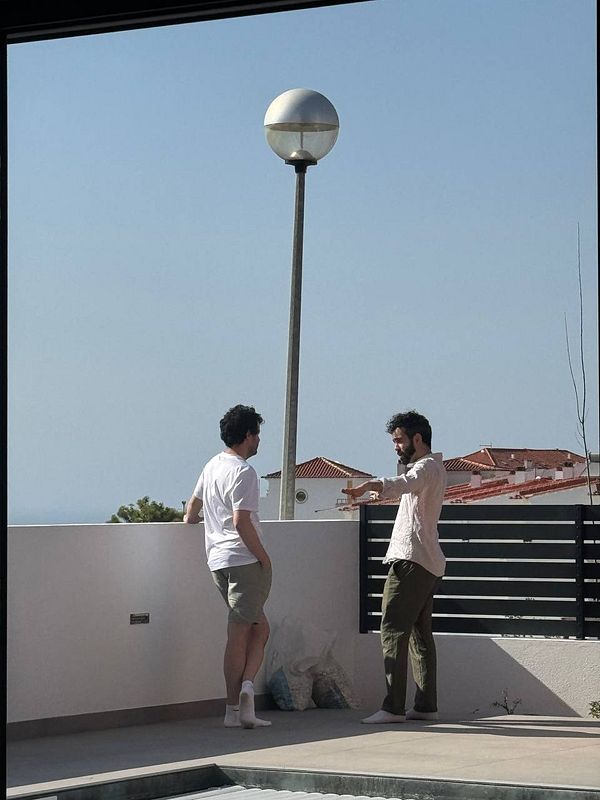 Two men are engaged in conversation on a balcony with a clear blue sky in the background.