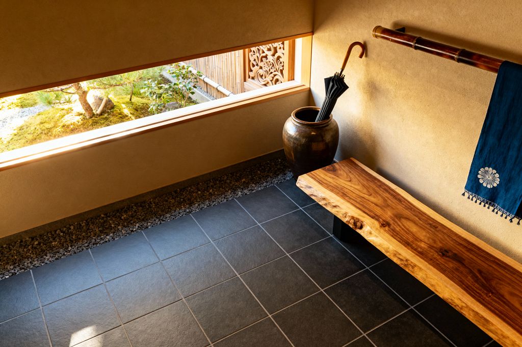 A serene Japanese-style mudroom with charcoal floor tiles and a border of pebbles features a live-edge wood bench with black legs against sand-colored plaster walls, a bamboo rail draped with an indigo cloth, and a large glazed ceramic jar holding umbrellas. A wide horizontal window frames a moss garden, stones, and a bamboo fence outside, bringing warm natural light into the space.