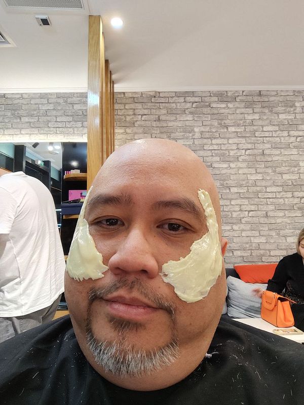 A man with a bald head and facial hair is sitting in a barber shop with a cream applied to his cheeks.