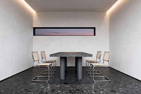 A minimalist dining room with a thick gray stone table on pedestal legs, surrounded by six chrome cantilever chairs with woven cane seats and backs. Smooth white plaster walls frame a narrow horizontal window showing a dusky sky, while warm cove lighting washes the ceiling. A dark charcoal terrazzo floor grounds the symmetrical, uncluttered arrangement, emphasizing clean lines and simple, tactile materials.