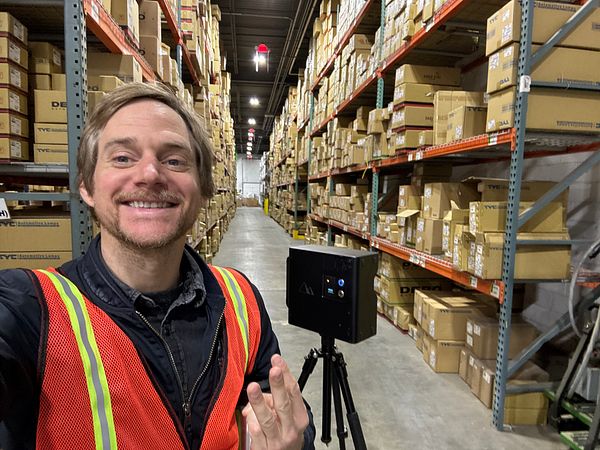 A person is smiling and posing for a selfie in a warehouse filled with stacked boxes.