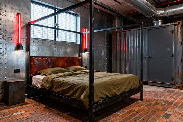 A raw industrial bedroom features a blackened steel four-poster bed with a cognac leather headboard and olive bedding on red brick flooring. Riveted sheet-metal walls frame a small window with wire mesh, flanked by vertical red neon tube sconces above matte black shades; a corrugated steel shipping-container door and exposed galvanized ductwork complete the warehouse aesthetic.