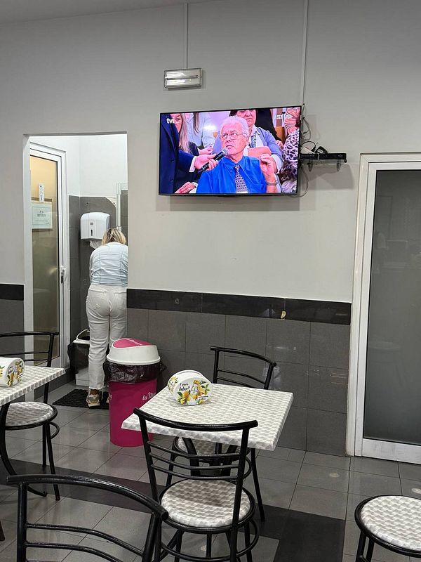 A small café area with a television showing a program, featuring a woman in white pants and a pink trash bin.