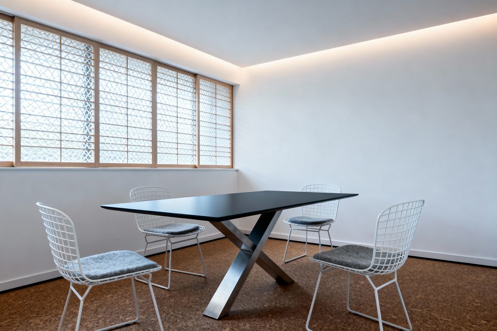 A minimalist dining room features a rectangular black table with an X-shaped brushed steel base, surrounded by four white wire chairs with gray felt cushions. Warm cork flooring contrasts with crisp white walls and a continuous cove of concealed LED lighting that washes the ceiling. A wall of windows with light natural wood frames and patterned translucent glass softly filters daylight, keeping the space bright yet private. The room is uncluttered, emphasizing clean lines and a calm, airy atmosphere.