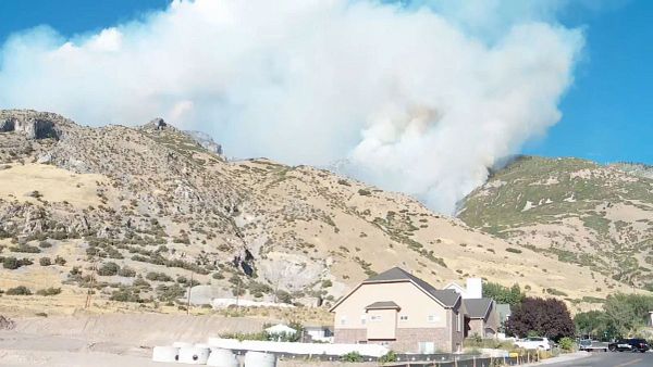 A large plume of smoke rises from a wildfire in a mountainous area near a residential building.