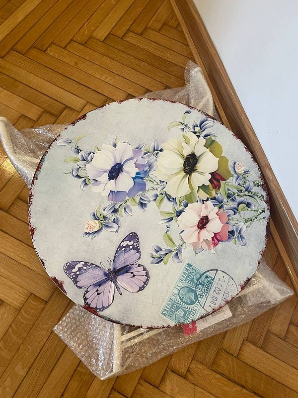 A decorative round table with floral designs and a butterfly is placed on a wooden floor.
