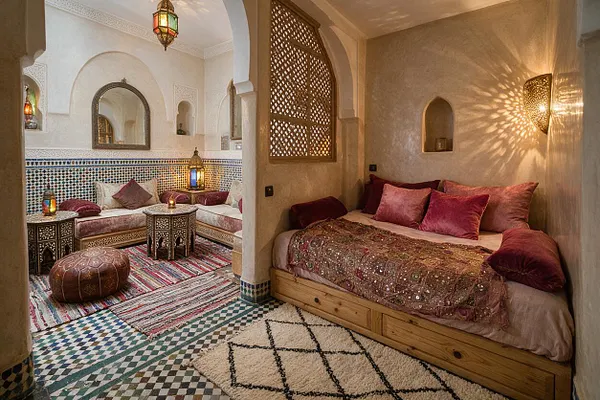 A Moroccan-style studio apartment with cream plaster walls, carved arches, and colorful mosaic tile details. The space includes a low wooden daybed with burgundy cushions, patterned rugs, ornate lanterns, and a small seating nook with a curved sofa and carved tables.