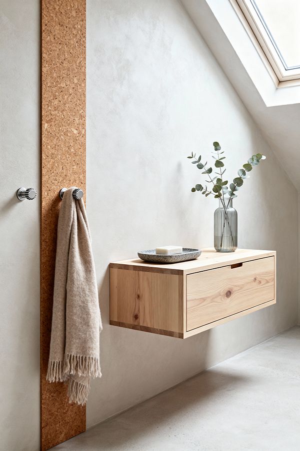 A minimalist Scandinavian mudroom beneath a skylight features warm off-white plaster walls and a vertical cork panel fitted with two knurled stainless-steel hooks, one holding a beige fringed wool scarf. A floating light-wood drawer cabinet with visible finger joints and a cutout pull projects from the wall, topped with a ribbed glass vase of eucalyptus and a speckled stoneware dish. The floor is smooth, light-gray concrete.