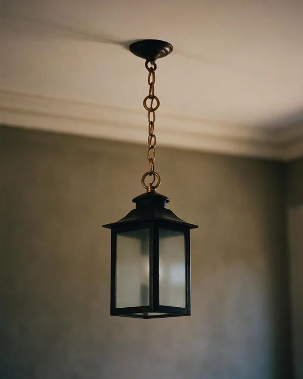 A hanging lantern-style pendant light with a black metal frame and frosted glass panels is suspended from the ceiling by a brass chain. It is centered against a neutral wall and ceiling, creating a simple, elegant Transitional look.