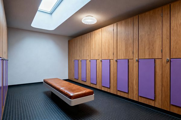 A streamlined mudroom features a long wall of vertical wood lockers with lower lilac doors and small chrome latches, a central bench with a cognac leather cushion on a slim pedestal base, and black rubber coin flooring. A large skylight and a white sculptural flush-mount light illuminate the warm wood and matte white walls, emphasizing clean mid-century lines and practical storage for shoes, bags, and outerwear.