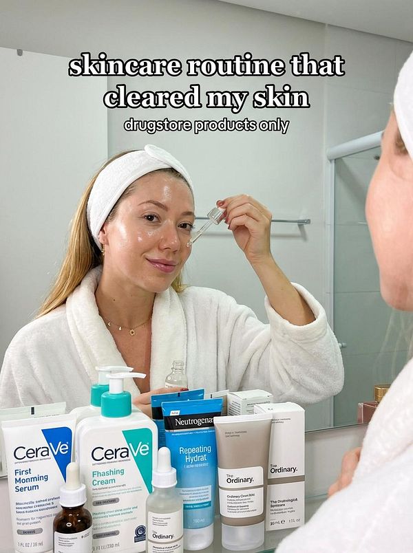 A woman demonstrates her skincare routine in a bathroom setting, surrounded by various drugstore skincare products.