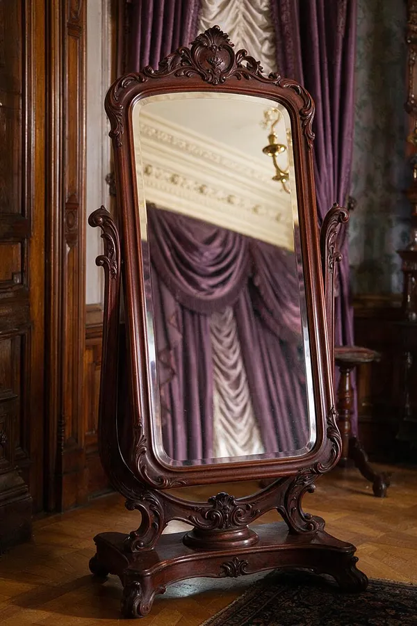 A tall Victorian-style floor mirror stands in a richly decorated room, featuring a carved dark wood frame with scrolling details and a shaped pedestal base. Purple drapery and paneled wood walls reflect in the mirror, reinforcing the formal, antique atmosphere.