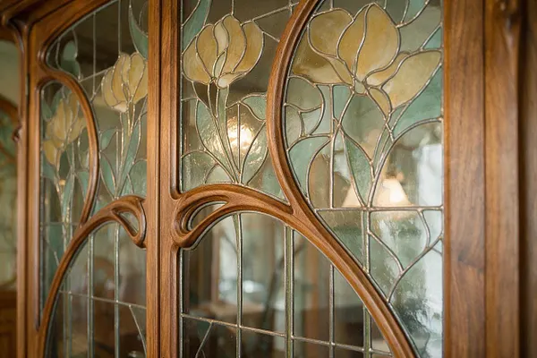 A close-up of an Art Nouveau wooden cabinet or door featuring curved carved wood frames and leaded glass panels. The glass is decorated with stylized floral designs in muted green, cream, and amber tones, with soft light glowing through the panes.