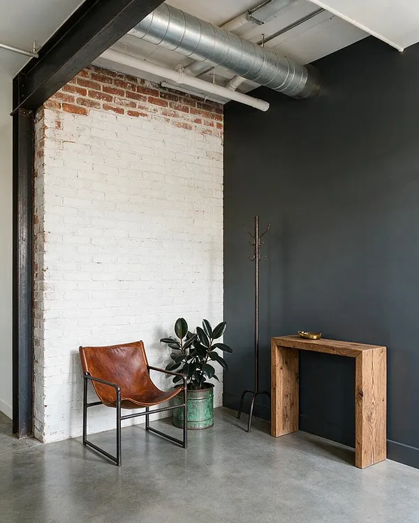 An industrial entryway with a whitewashed exposed brick wall, dark charcoal walls, and visible ceiling ductwork and pipes. A leather sling chair, potted plant, slim metal coat rack, and rustic wood console create a simple, urban welcome area.