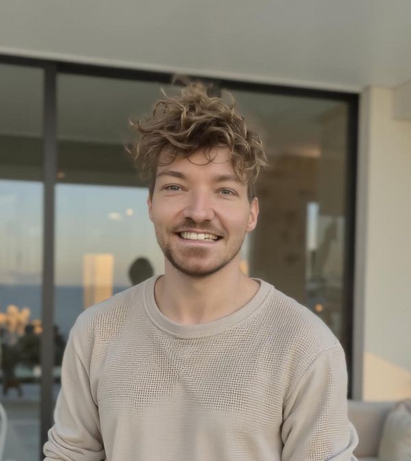 A smiling young man with curly hair stands in front of a large window, showcasing a modern interior.