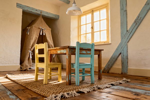 A cozy farmhouse-style kids’ room features a small weathered wooden table with two child-size chairs, one aqua and one butter yellow, set on a chunky jute rug over wide plank floors. Sunlight pours through a pale yellow, multi-pane window. A burlap play tent with tied openings nestles in the corner, while distressed duck-egg blue timber beams cross the cream plaster walls and a white enamel pendant light hangs overhead, creating a warm, rustic play space.