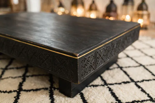 A low rectangular Moroccan-style coffee table with a dark carved base and smooth black top, outlined by a thin brass trim. It sits on a cream rug with black geometric lines, with softly glowing lanterns blurred in the background.