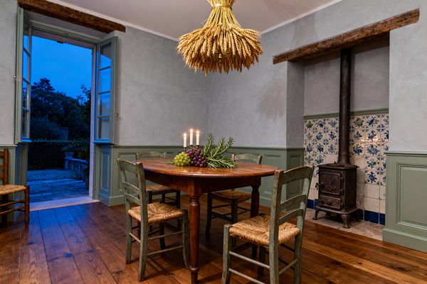 A French Country dining room centers on a round wooden table surrounded by four distressed green ladder-back chairs with rush seats, topped with candles, grapes, and rosemary beneath a wheat-sheaf pendant. Sage green wainscoting and window trim frame pale gray plaster walls, while an antique cast-iron stove stands in a blue-and-white tiled alcove under a timber beam, with wide-plank floors leading to open French doors and an evening garden.