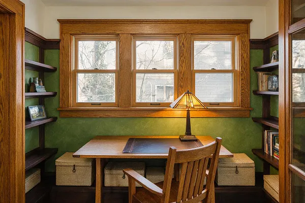 A Craftsman-style sunroom features a central wooden desk beneath three large wood-trimmed windows, with a slatted wooden chair and a warm table lamp. Built-in dark wood shelves line both sides of the room, while green walls and woven storage baskets create a cozy, functional nook filled with natural light.