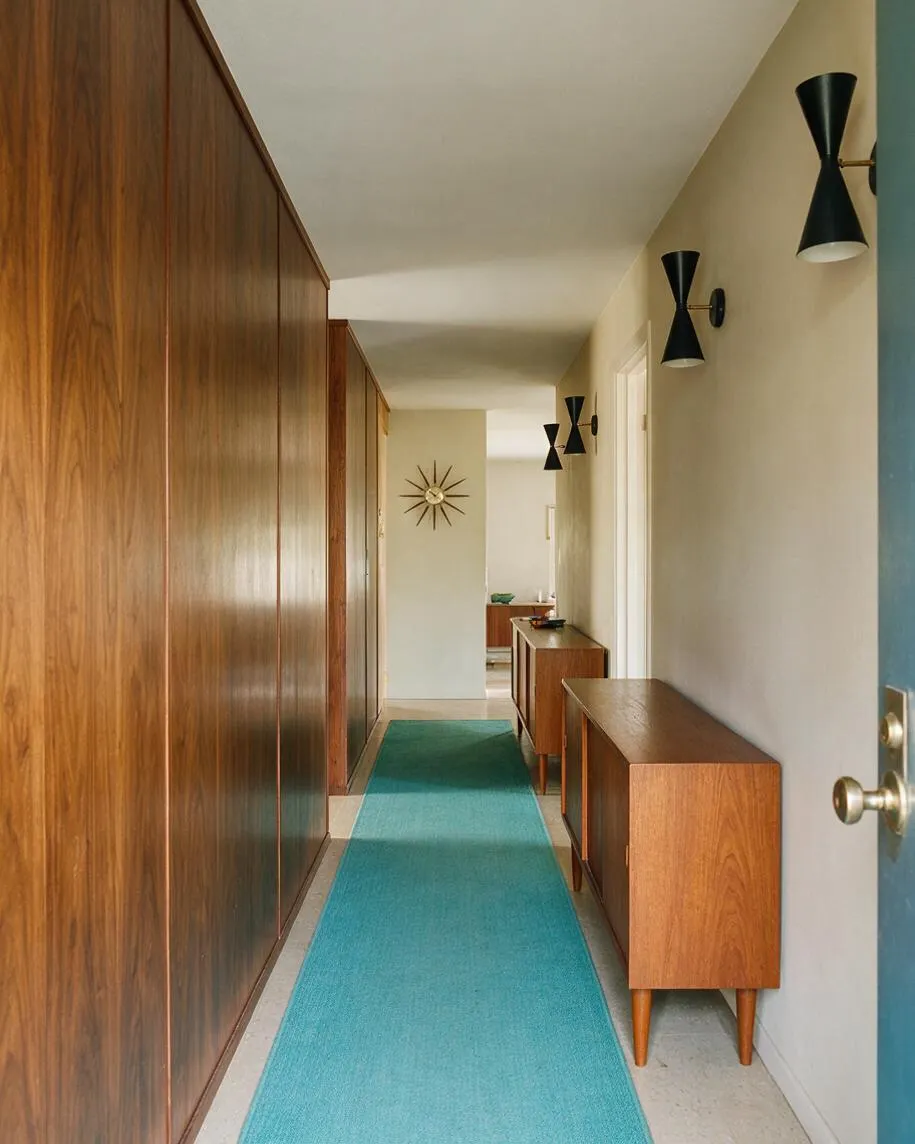 A long, narrow entryway with warm walnut wood paneling and storage on the left, a turquoise runner rug on the floor, and beige walls on the right. Black sconce lights, a brass starburst wall clock, and mid-century wooden cabinets create a clean, retro-inspired look.